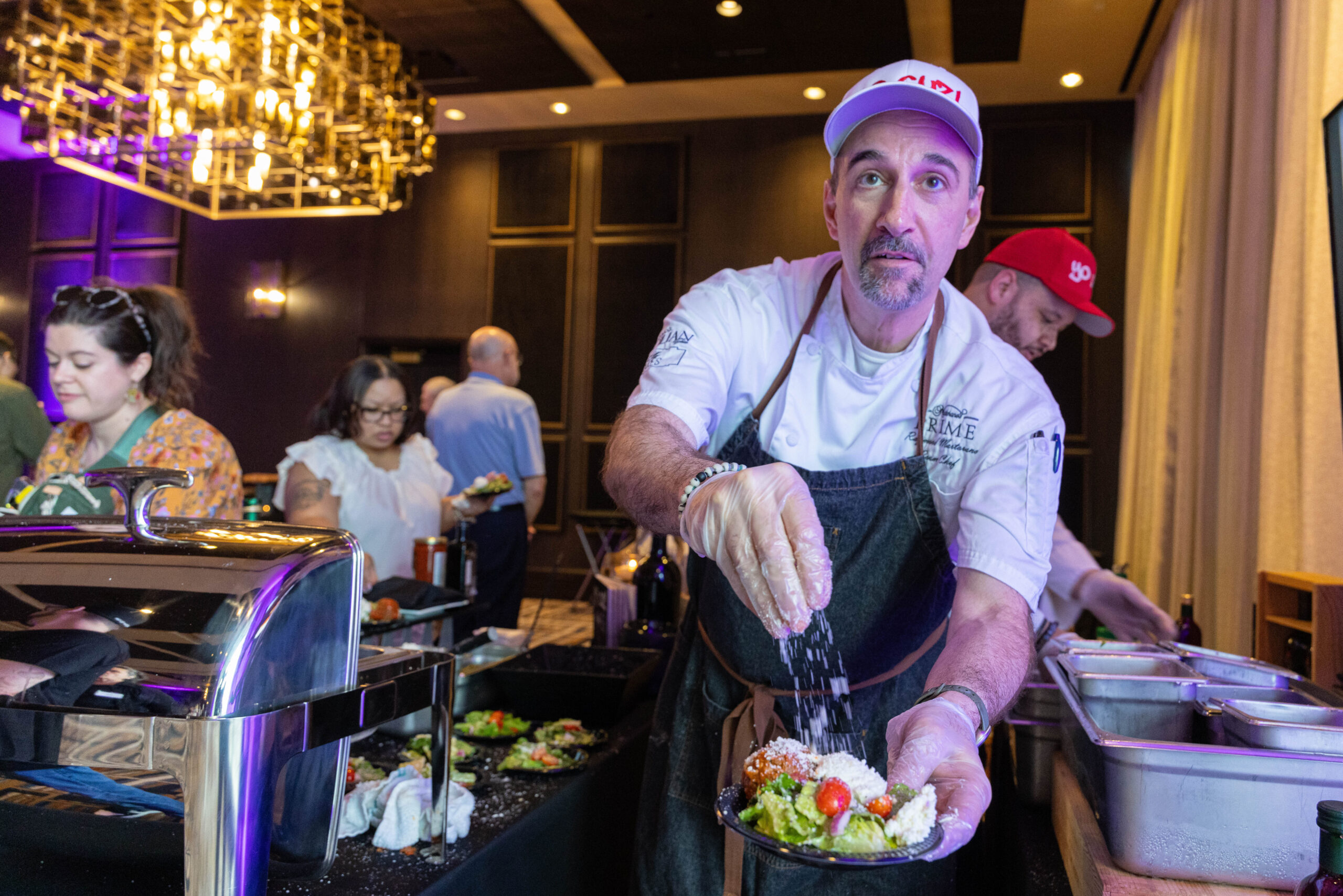 A chef sprinkles cheese on a salad while looking at the camera